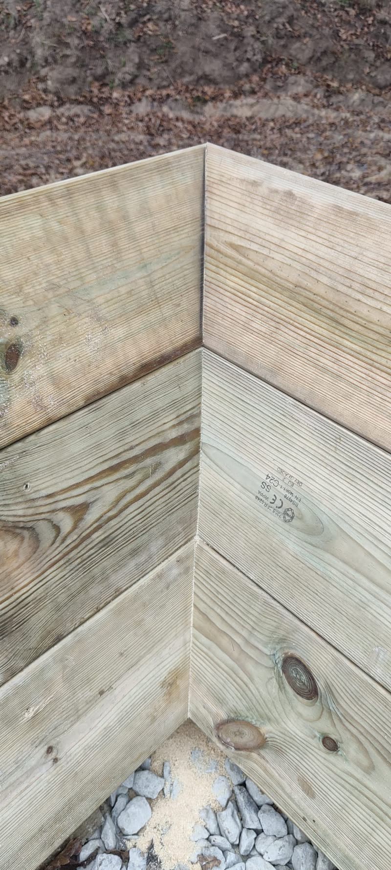A timber kickboard corner joint, boards mitred flush, stone and sand visible below