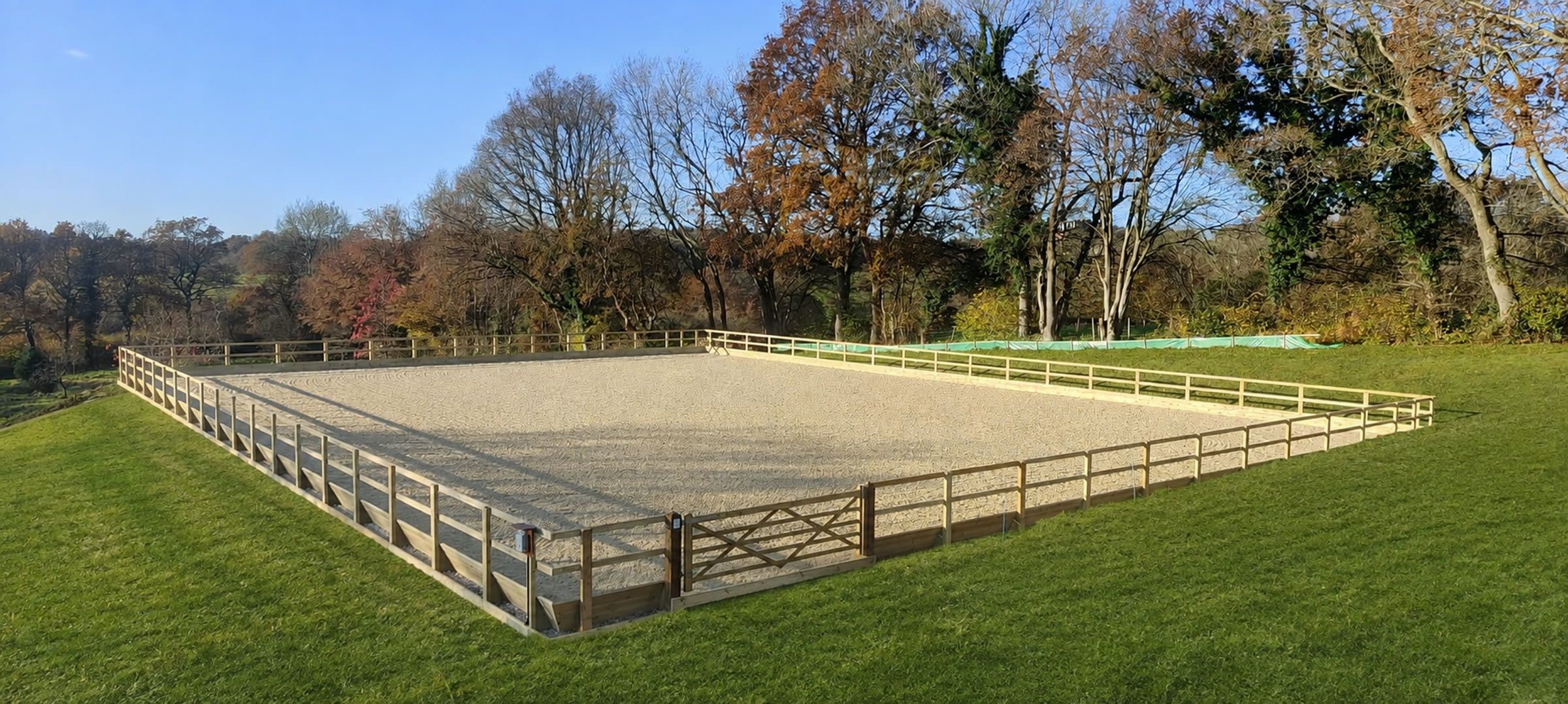 A completed riding arena with a levelled sand surface, timber kickboards and post and rail fence, looking out over wooded countryside